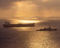A naval destroyer escorts a large cargo tanker silhouetted against a golden sunset