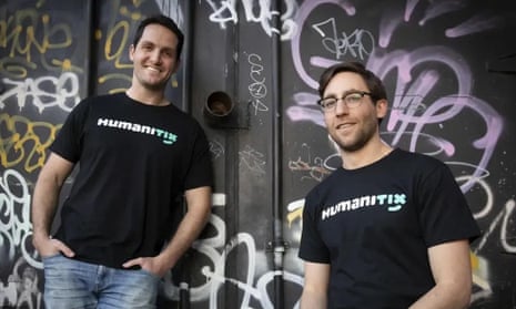 Adam McCurdie (left) and Josh Ross, founders of Humanitix, wearing black T-shirts bearing the Humanitix name. They are standing in front of a black wall that is covered in graffiti.