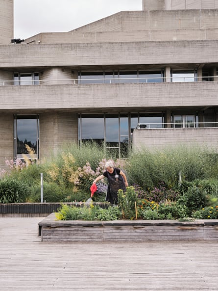 a woman watering the dye garden with the brutalist theatre building behind her