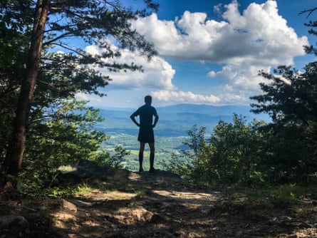 A person stands in a forest overlooking mountains and a valley.