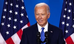 Man half-smiles in front of microphone with American flag in background