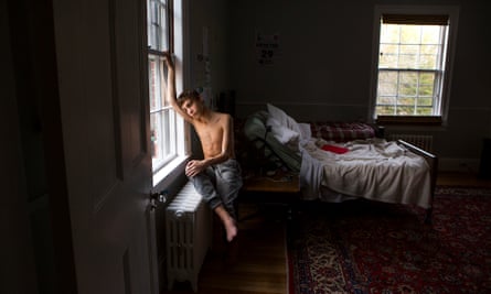 A boy who is naked from the waist up sits on a radiator as he gazes out of his bedroom window, Two beds are in the background