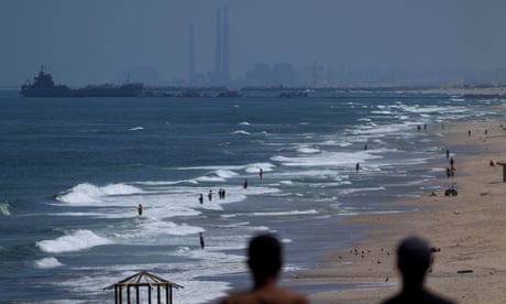 A ship is anchored next to a pier on which vehicles can be seen, at a surf beach with people standing on the water's edge