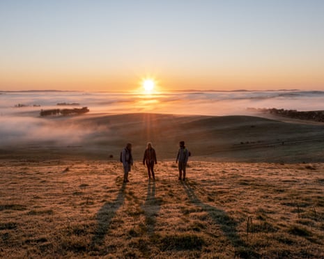 Three hikers watch the sun rise out of the mist shrouding dry paddocks