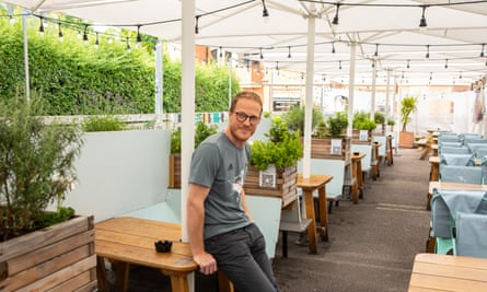 Ed Whinney, in a T-shirt and jeans, leaning against one of a number of outdoor tables under canopies at his pub