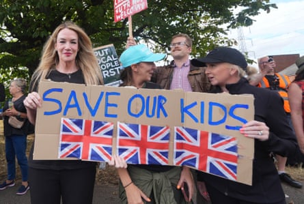 Three women hold up a cardboard sign with Save Our Kids written on it and three union jack flags. More people with placards stand in a crowd behind them.