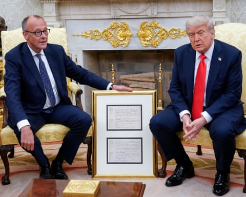 Merz (left) and Trump sitting in the Oval Office as Merz rests his hand on the framed birth certificate as he holds it on the floor