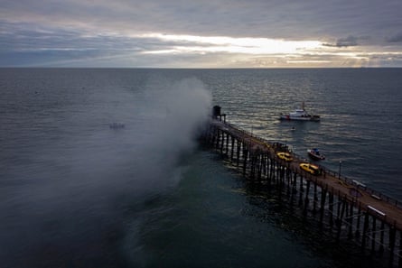an aerial view of a long wooden structure extending into the ocean with smoke rising from the end