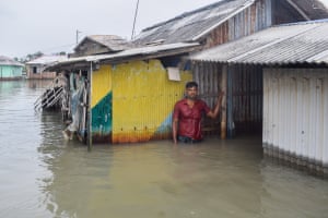 A man stands up to his waist in floodwater by a house made of sheets of corrugated iron.