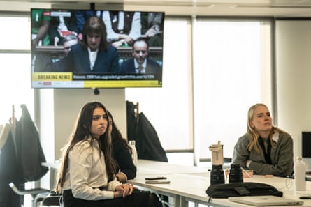 three young women, two with long dark hair and one blond, sit at a long table watching a screen; another TV screen is on the wall behind them showing Rachel Reeves giving her speech.