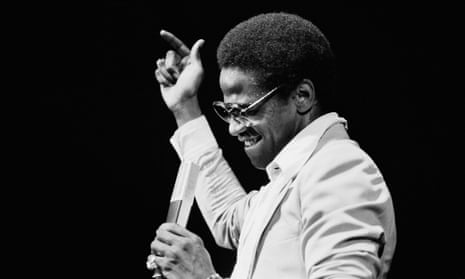 Black and white image of American singer Al Green performing on stage. The image shows his head and shoulders. He is holding a microphone, and his other hand is above his head