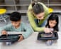 A teacher points to a tablet screen as two children view their own tablets in class