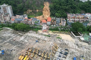 An aerial view shows several collapsed buildings in the aftermath of a mudslide down the tree-lined hillside behind them; in the foreground, yellow diggers and construction vehicles are lined up