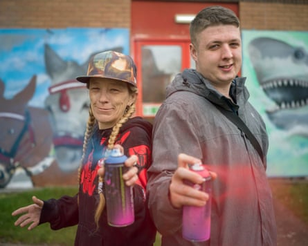 A woman in a cap and a man stand back to back deploying spray cans towards the camera