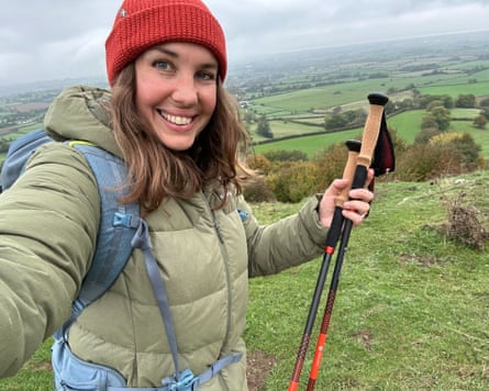 A smiling woman, writer Sian Lewis, in a red beanie holds trekking poles overlooking green countryside