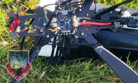 Part of a drone, with a circuit board and wires coming out of it, on a patch of grass