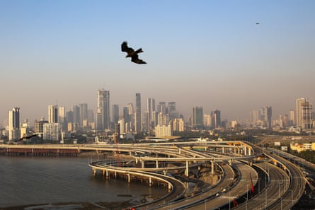 A view of a multi-lane motorway on piles with slip roads curling off it amid a smoggy cityscape with tower blocks on the horizon