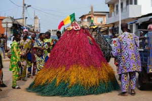 A person in Vodún attire, dressed as Zangbeto, wearing a large raffia costume in the shape of a haystack, coloured in single bands of green, yellow and red