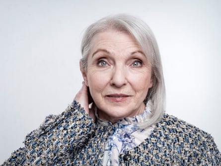 A woman with short grey blond hair, wearing a black speckled jacket and a blue printed shirt.