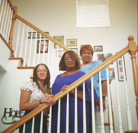 Three women stand on a flight of stairs in a home. The walls behind them are filled with family pictures.