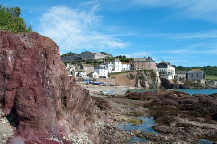 A small red sandstone rock cliff beside a rocky beach opening up to the village of Cawsand.