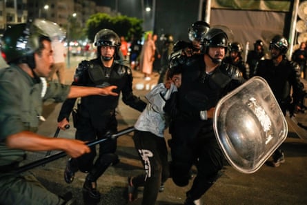 A scared0-looking boy is dragged away by a riot policeman in a helmet and carrying a shield as other police run alongside