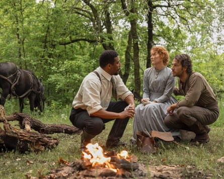 Man squats while talking to man and woman in woods by campfire