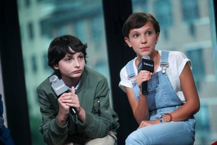 Wolfhard wearing a green jacket and Brown wearing a white T-shirt and light blue denim dungarees, holding up microphones