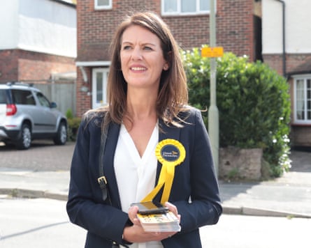 Monica Harding campaigning in Surrey. She is about 50 and has long, straight brown hair; she wears a dark jacket over a white blouse, with a large yellow Lib Dem rosette on her collar.