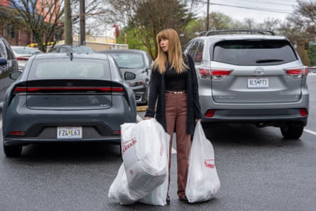 Linda Cardellini carrying shopping bags stands between parked cars on a wet street, looking anxious
