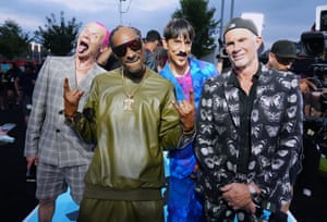 Flea, Snoop Dogg, Anthony Kiedis and Chad Smith pose for a photo. Flea has bright pink hair and has his tongue out. Snoop is wearing a green leather outfit and sunglasses. Kiedis is wearing a bright blue and pink suit, and Smith wears a navy blazer with white butterflies on it and a backwards cap