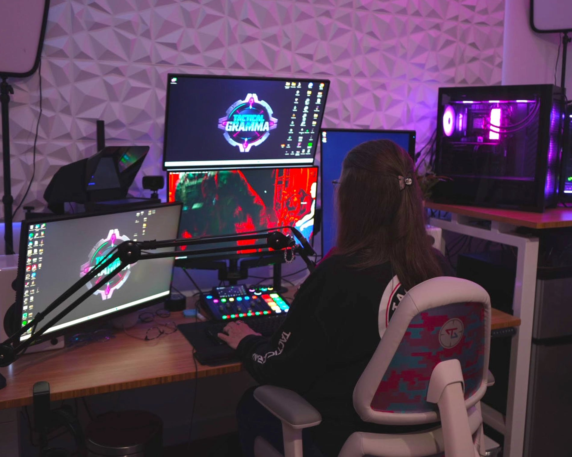 Mujer sentada frente a un completo setup de streaming con múltiples monitores, micrófono, consola de audio y luces LED moradas, en una habitación decorada con paneles acústicos.