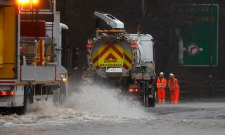 Emergency vehicle pumping water on flooded road with two workers in high vis workwear standing beside them.