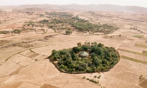 A round church roof is seen amid trees surrounded by dusty beige fields