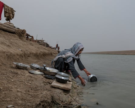 A woman crouches on the shore next to piles of tin dishes and pans, dipping a pot into the murky water in front of her