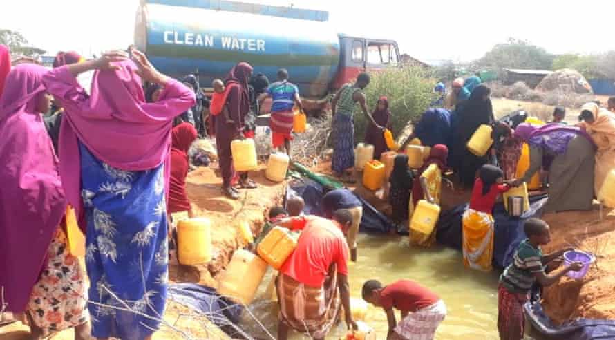 Somali people fill containers from a temporary pool of water. A truck with 'clean water' written on it is in the background.