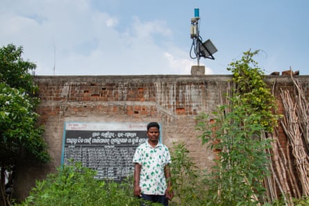 A man stands in front of a brick wall with a blackboard on it with writing in a local script. An aerial with equipment on it stands on top of the wall