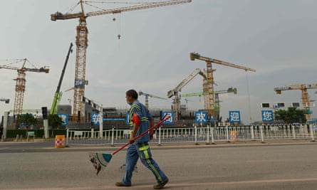 A man in work clothes carrying a rake walks in front of a series of cranes
