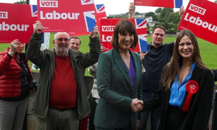 Rachel Reeves shakes hands with Jade Botterill as they stand in front of a group of men holding red Vote Labour placards. Reeves wears a dark green jacket with purple silky shirt and Botterill wears a black jacket with blue shirt; she has a large red rosette on her lapel.