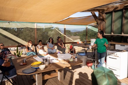 A group of people sit around a table and under a shade outdoors, on a wooden deck.