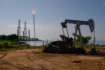 An oil pumpjack beside a lake on a sandy shore, with a flare and tall iron structures on the background against a blue sky.