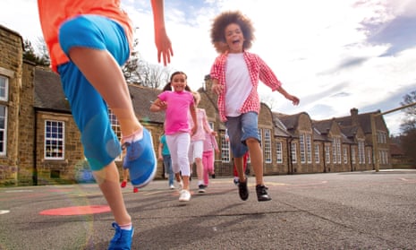 Children running across the school playground smiling. The children are running towards the camera. There are motion blurs and a sun flare. The children are casually dressed for summer. The school is visible in the background.