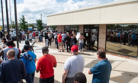 A group of people wearing mostly T-shirts and baseball camps stand in a crowded line that snakes into a circle outside a one-story beige building with floor-to-ceiling windows.