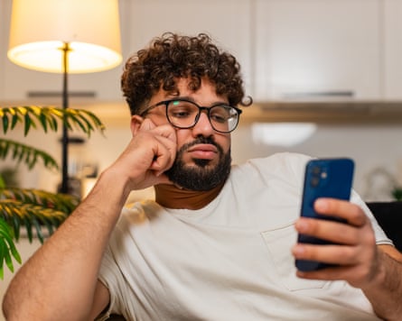 A dark haired man with glasses looks bored as she scrolls on his phone.