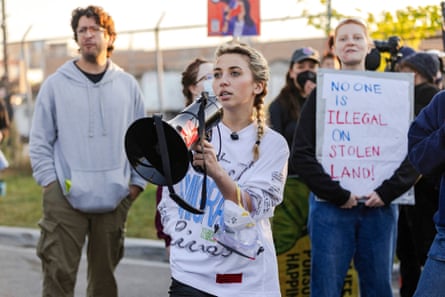 A woman with a bullhorn, with protesters behind her, along a chain-link fence with barbed wire.