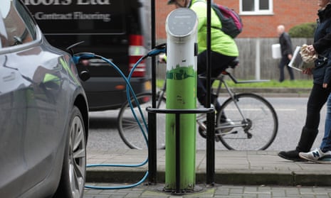Pedestrians and a cyclist pass a car plugged into a charging point