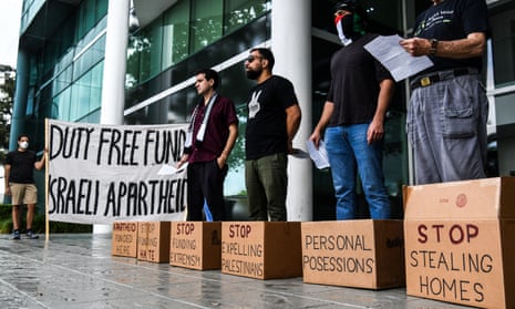 Four men stand behind cardboard boxes with protest messages on them that are at their feet. Another man stands nearby holding a large sign that reads 'Duty free fund Israeli apartheid'.