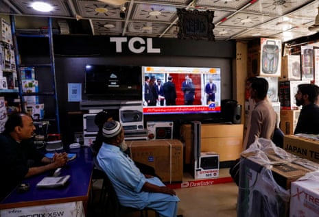 Five men in a white goods shop watch the news on TV showing JD Vance's arrival in Islamabad.