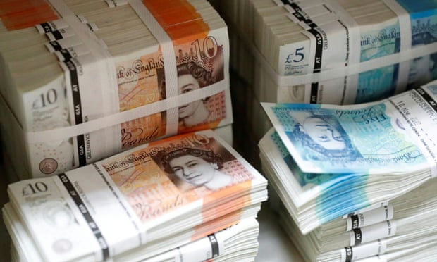 Wads of British Pound Sterling banknotes are stacked in piles.