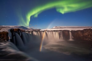 The image is taken from the edge of a waterfall which curves around, lined by snow-covered rick. A rainbow appears to be rising from the water at the bottom of the waterfall and into the dark blue sky. A streak of bright green light is in the shape of a wave across the sky